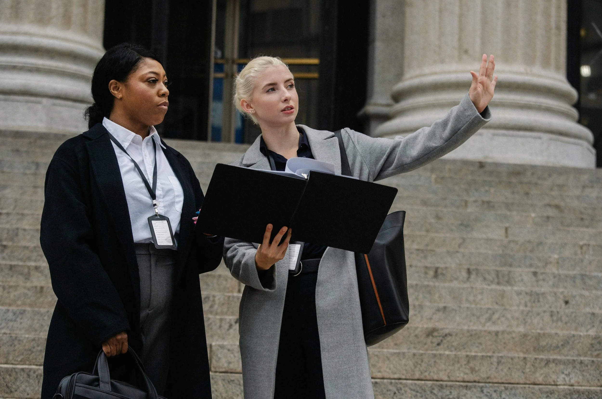 Two women standing on steps outside a building, engaged in conversation. One woman is holding a folder and gesturing, while the other woman listens.