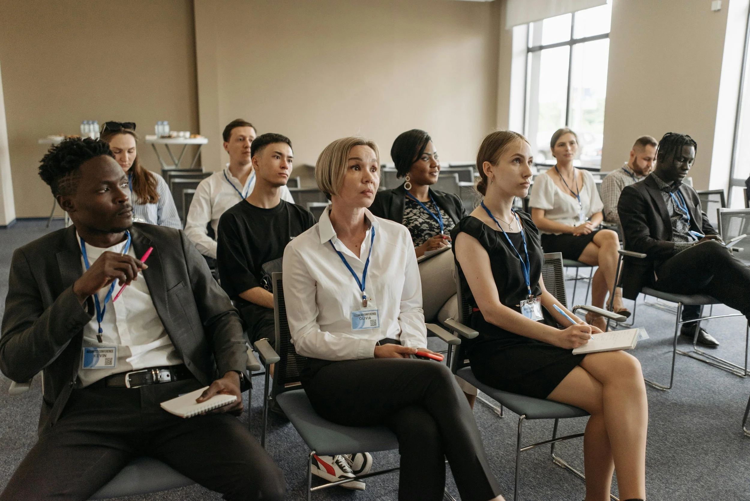 A group of diverse people attending a conference or seminar, sitting in chairs in a room with large windows.