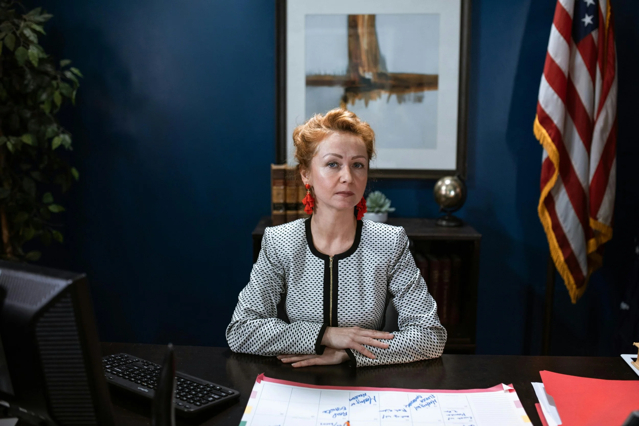 A woman with red hair and red earrings sitting at a desk in an office, with an American flag, a painting, and office supplies in the background.
