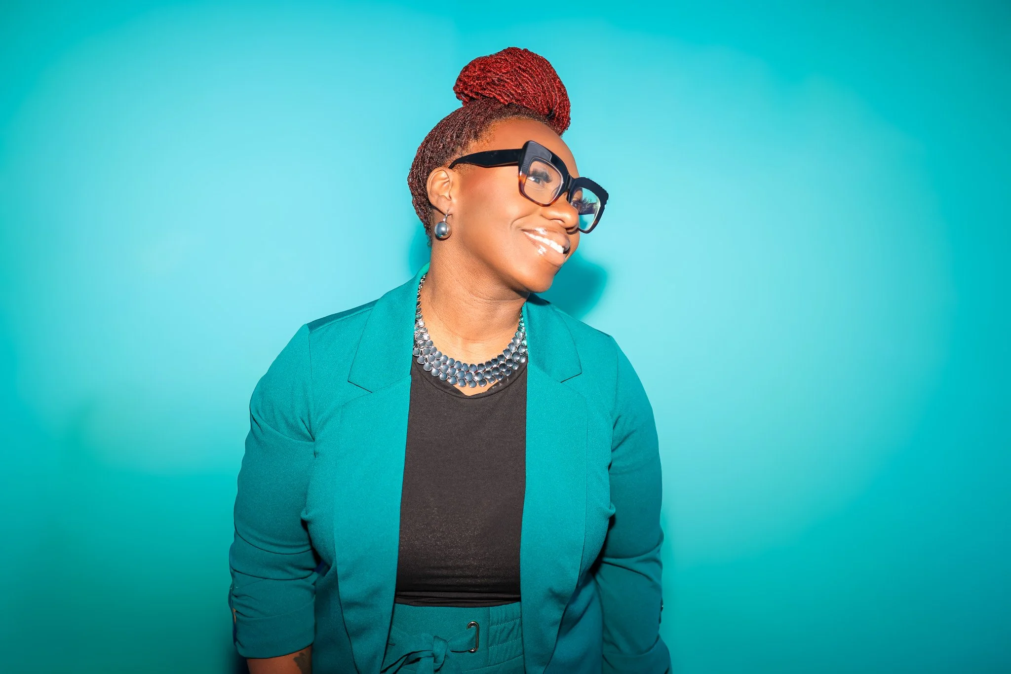 A woman with red braided hair, wearing large black glasses, a teal blazer, and a black top, smiling and looking to the side against a teal background.