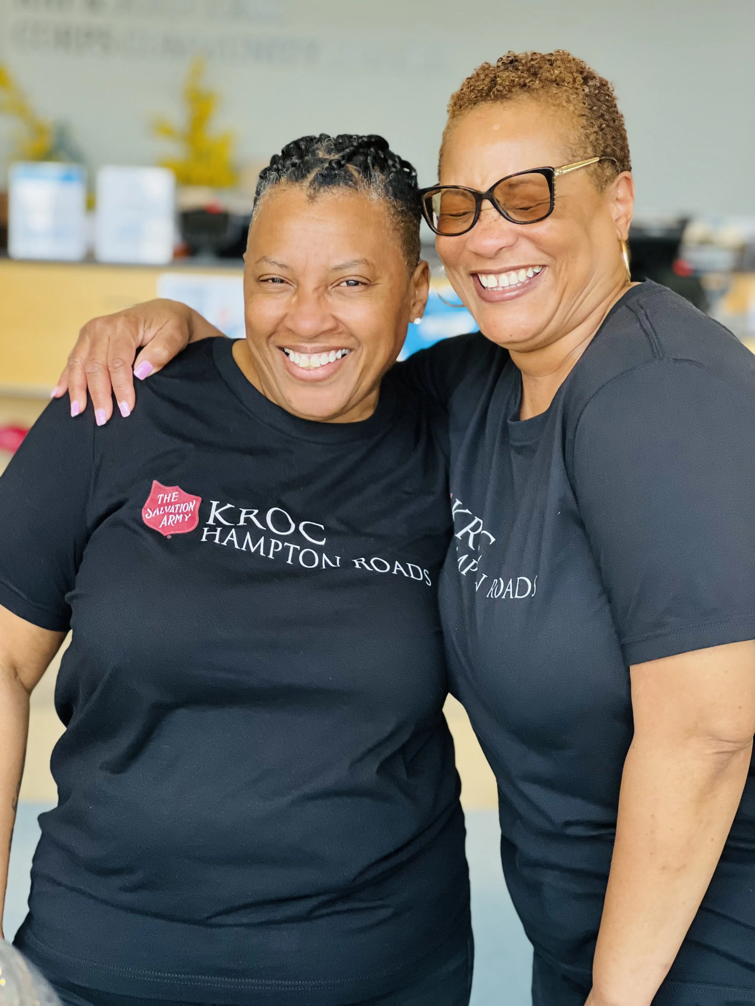 Two women smiling and hugging, wearing black T-shirts with 'KROC HAMPTON ROADS' and 'THE SALVATION ARMY' logos, indoors.