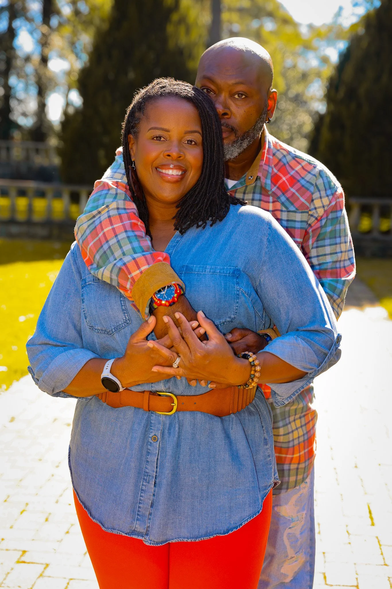A smiling woman with dark hair and a man with a beard hugging her from behind, outdoors on a sunny day with trees in the background.