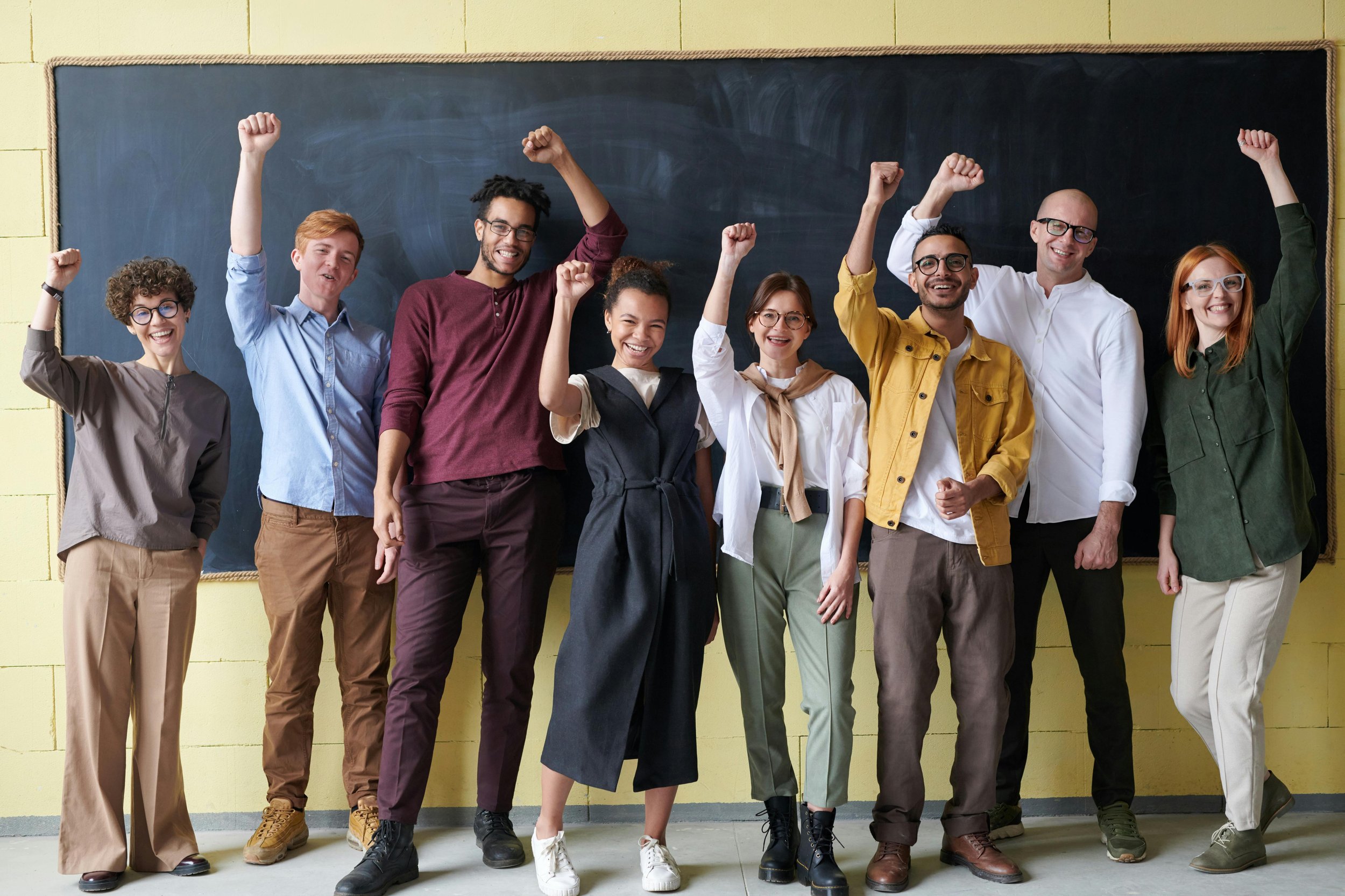 Group of diverse young adults celebrating with raised fists in front of a blackboard.