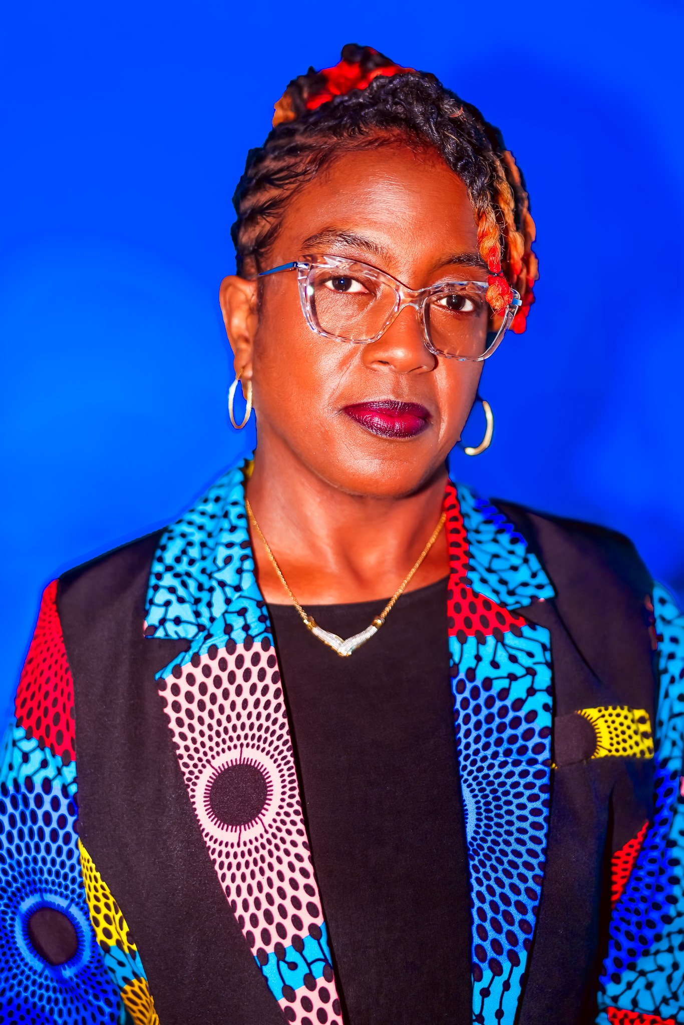Portrait of an African American woman with glasses, wearing vibrant, patterned clothing and gold jewelry against a blue background.