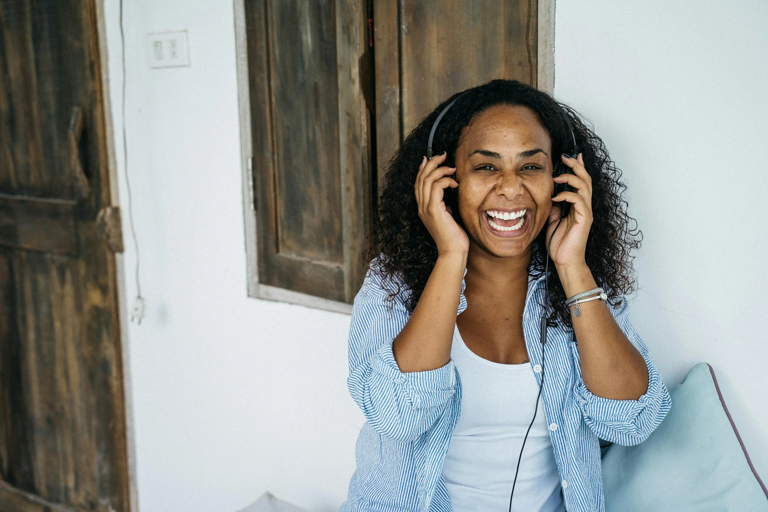 A woman with curly hair smiling while wearing headphones, sitting on a light-colored couch against a white wall with wooden window shutters.
