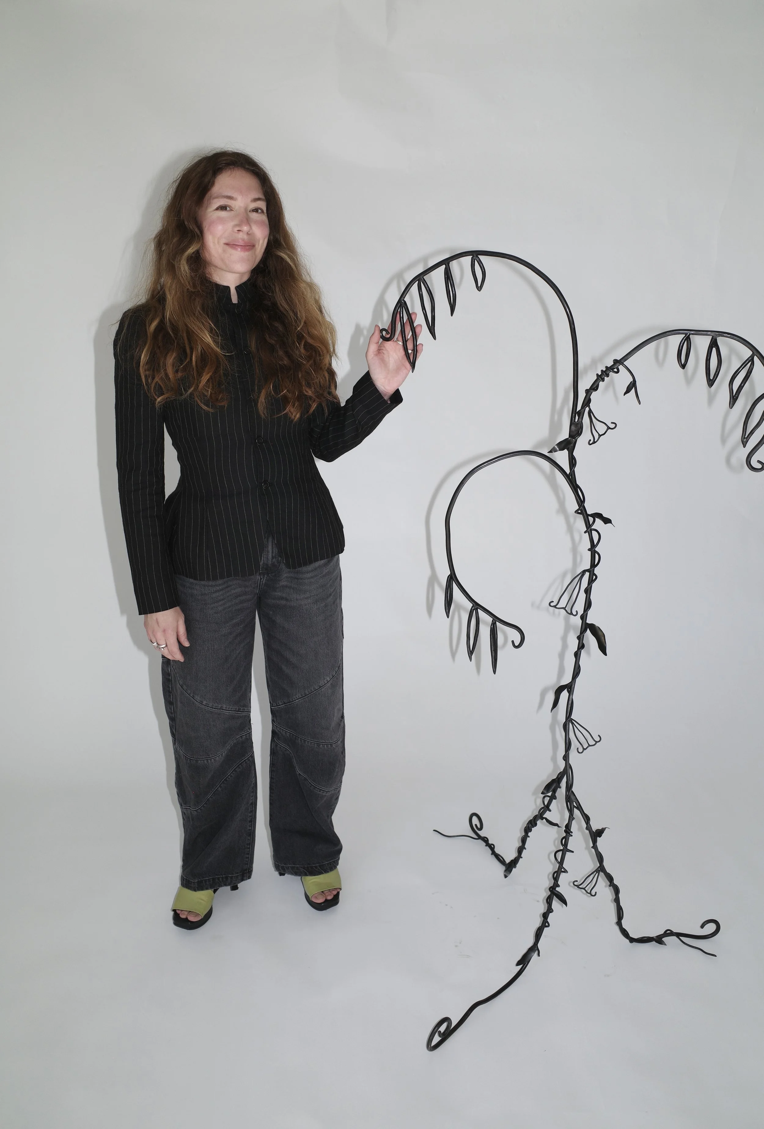 Woman with long curly hair wearing a black pinstripe blazer and black jeans standing next to a large black wrought iron sculpture resembling plant vines.