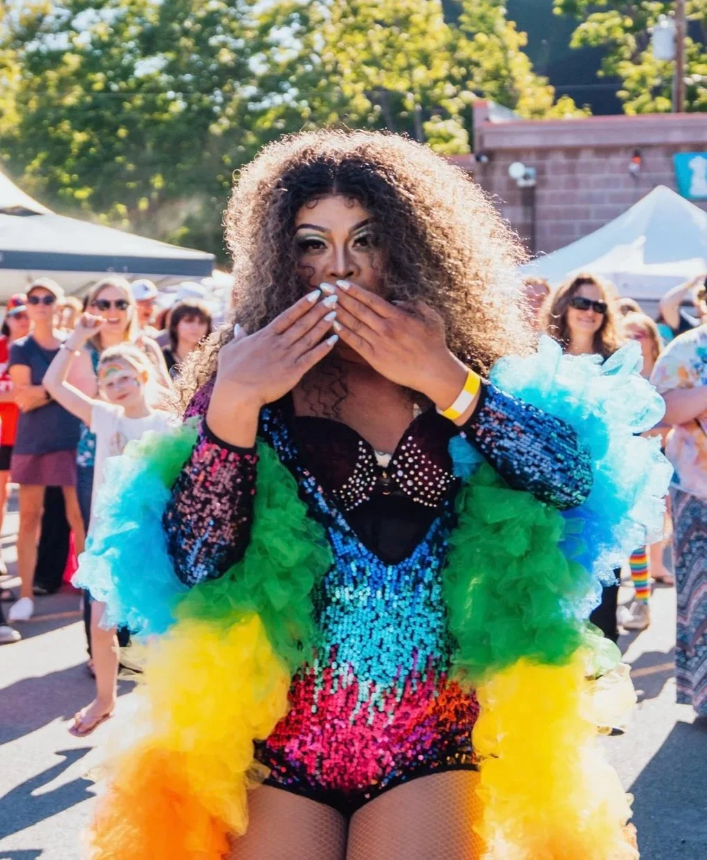 A drag queen in sequins and a rainbow boa prepares to blow a kiss. She looks fabulous.