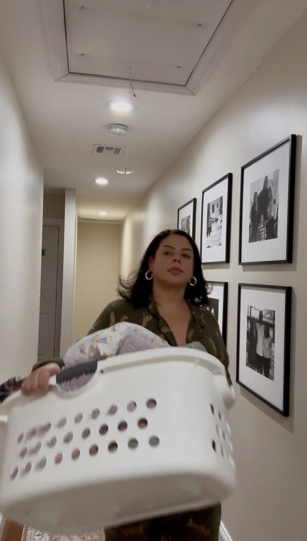 Michelle wearing gold earrings and a patterned shirt is walking down a hallway while carrying a laundry basket. The hallway has framed black-and-white photographs on the walls and ceiling lights.