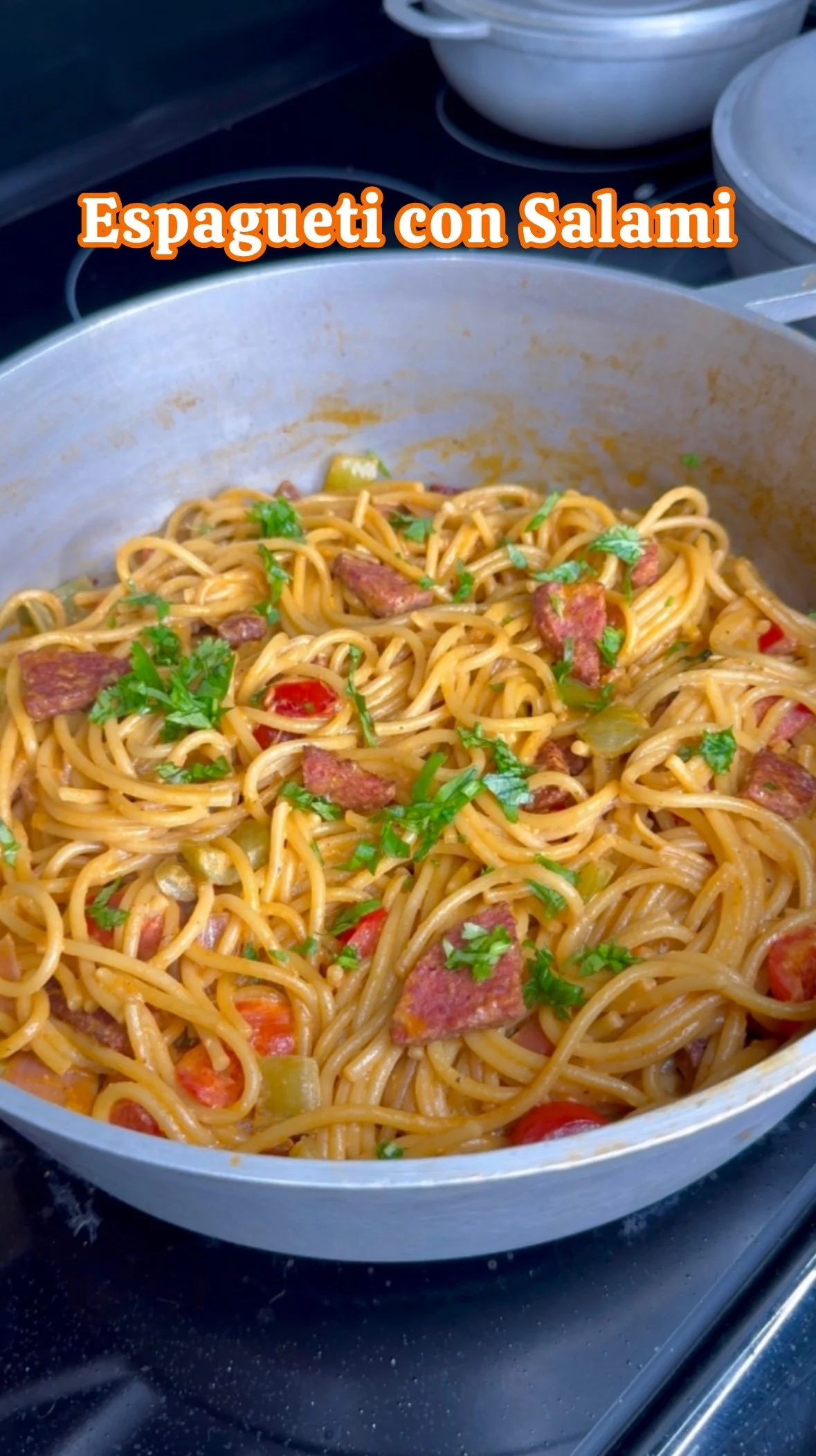 A pot of spaghetti with salami, cherry tomatoes, and herbs, labeled in Spanish as 'Espagueti con Salami'.