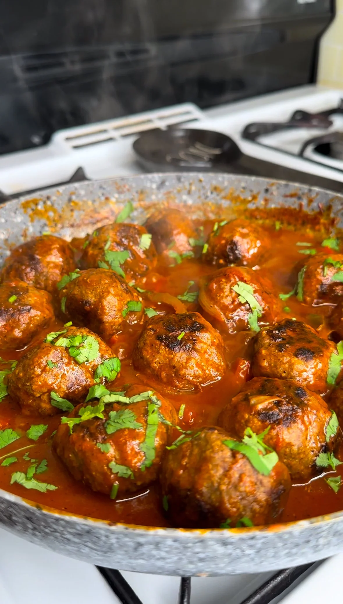 Cooked meatballs simmering in tomato sauce garnished with fresh cilantro in a pan on a stove.
