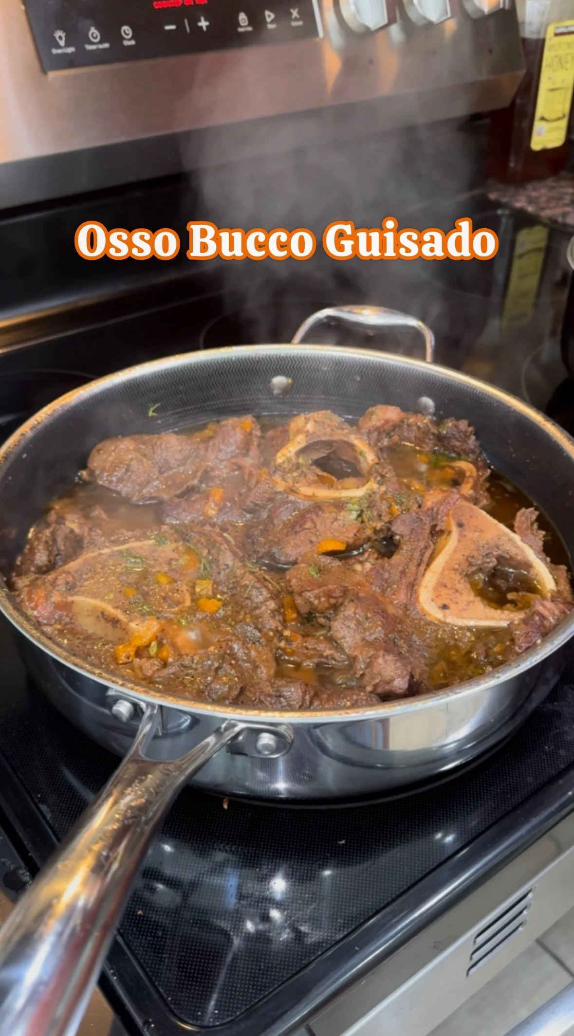 A stainless steel frying pan on a stovetop contains cooked beef shanks with broth and vegetables, emitting steam. The image includes a caption that reads "Osso Bucco Guisado" in orange and white text.