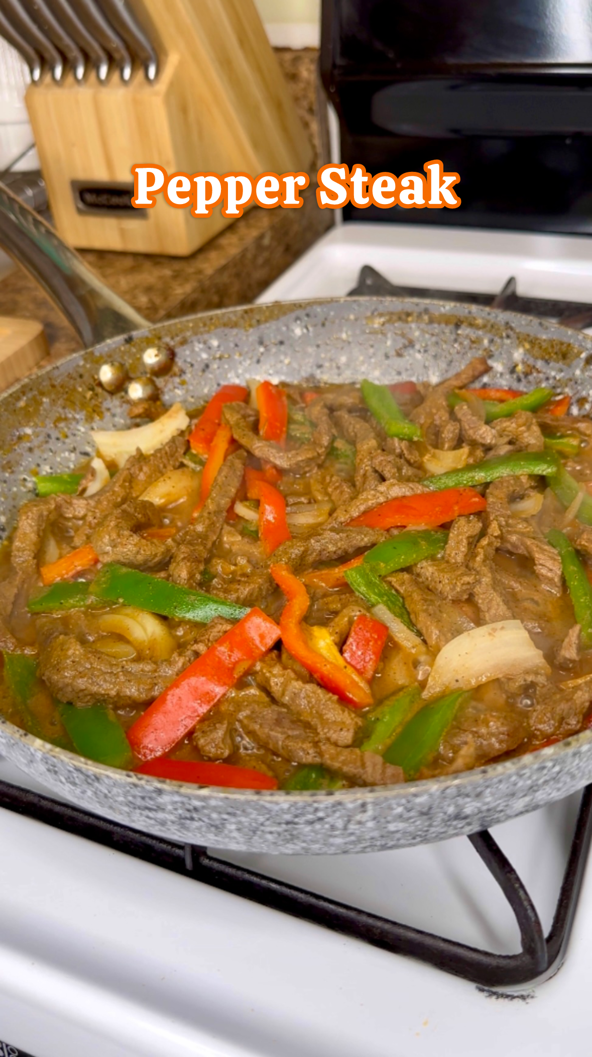 A skillet with cooked pepper steak, including red and green bell peppers and onions, on a stove with a wooden knife block in the background.
