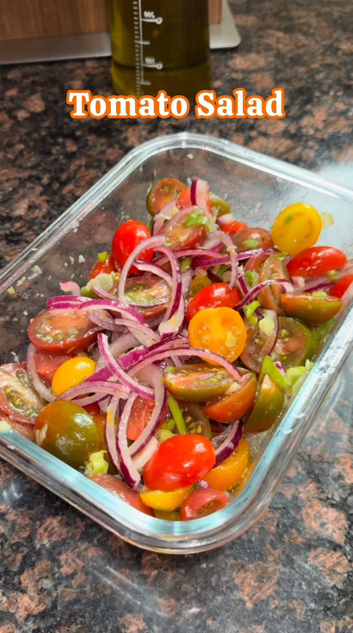A rectangular glass dish containing a fresh tomato salad with halved grape tomatoes and sliced red onions, placed on a dark granite countertop.