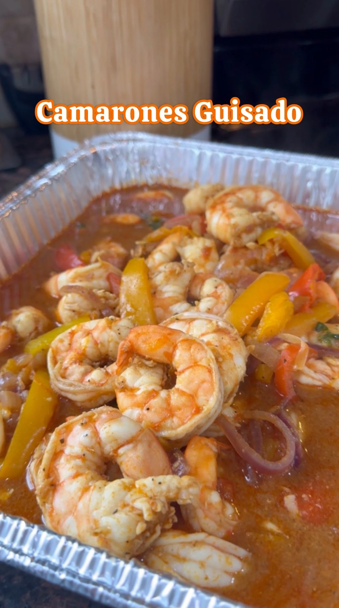 A container of cooked shrimp with yellow and red peppers in a savory sauce, with the text 'Camarones Guisado' overlaid.
