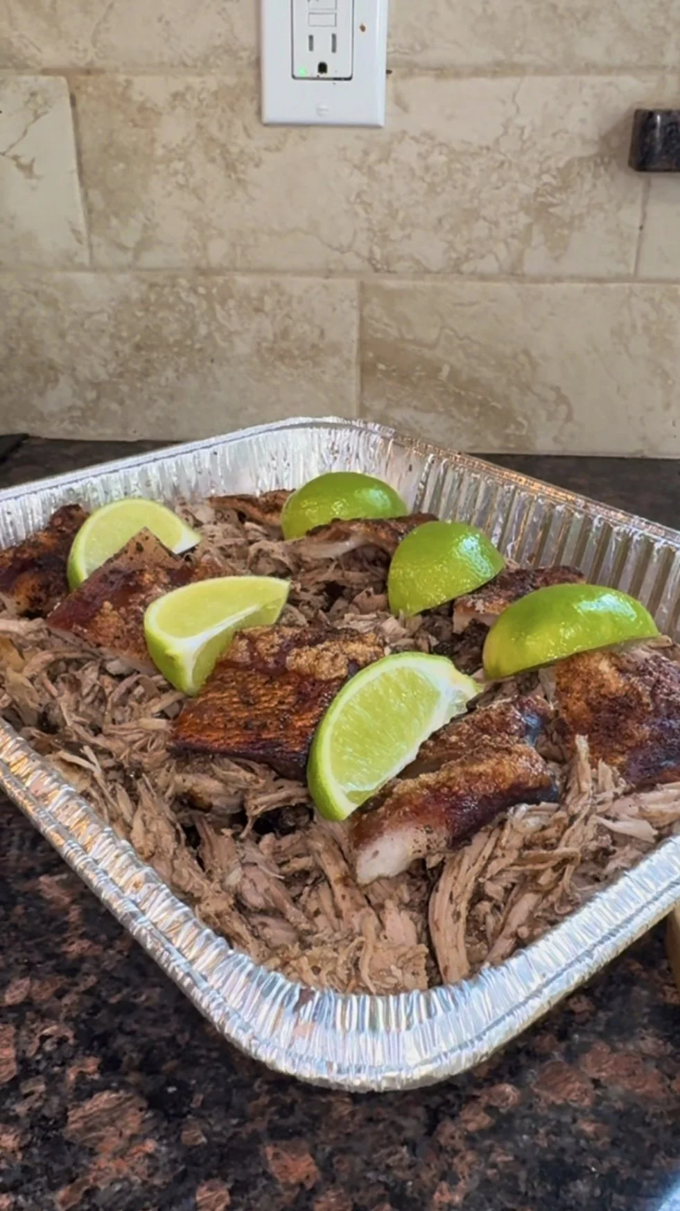 baked pork shoulder (pernil), garnished with lime wedges, in an aluminum tray on a kitchen countertop.