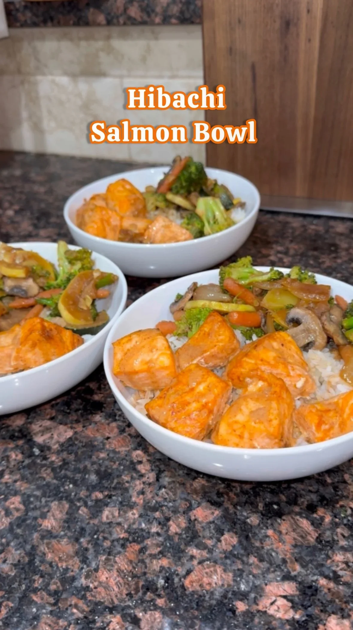 Three white bowls filled with rice, cooked salmon, and mixed vegetables including broccoli, carrots, and mushrooms, on a dark marble countertop, with the label "Hibachi Salmon Bowl" in orange text.