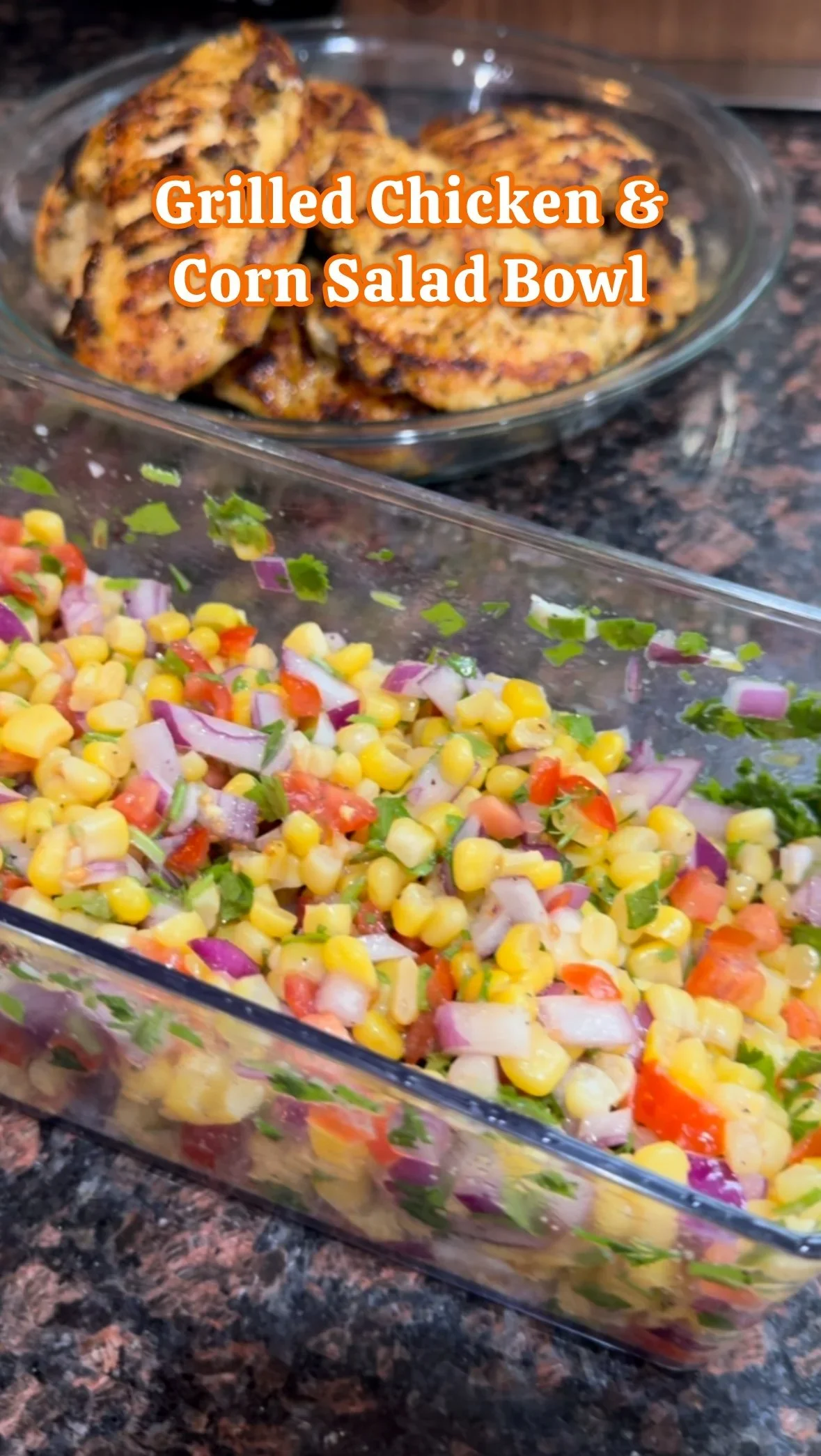 A glass dish of corn salad with white rice, chopped red onions, tomatoes, cilantro, and corn kernels in the foreground, and grilled chicken pieces in the background.