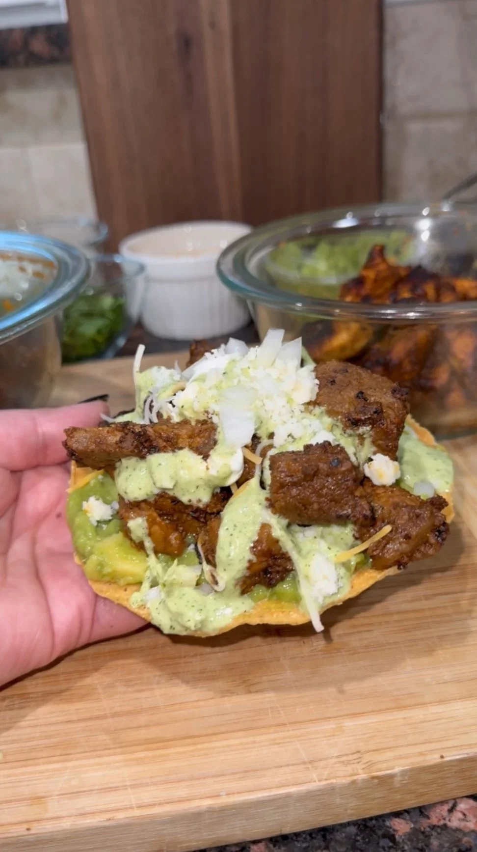 Close-up of a tostada with grilled steak, shredded cheese, lettuce, guacamole, and sour cream on a wooden cutting board.