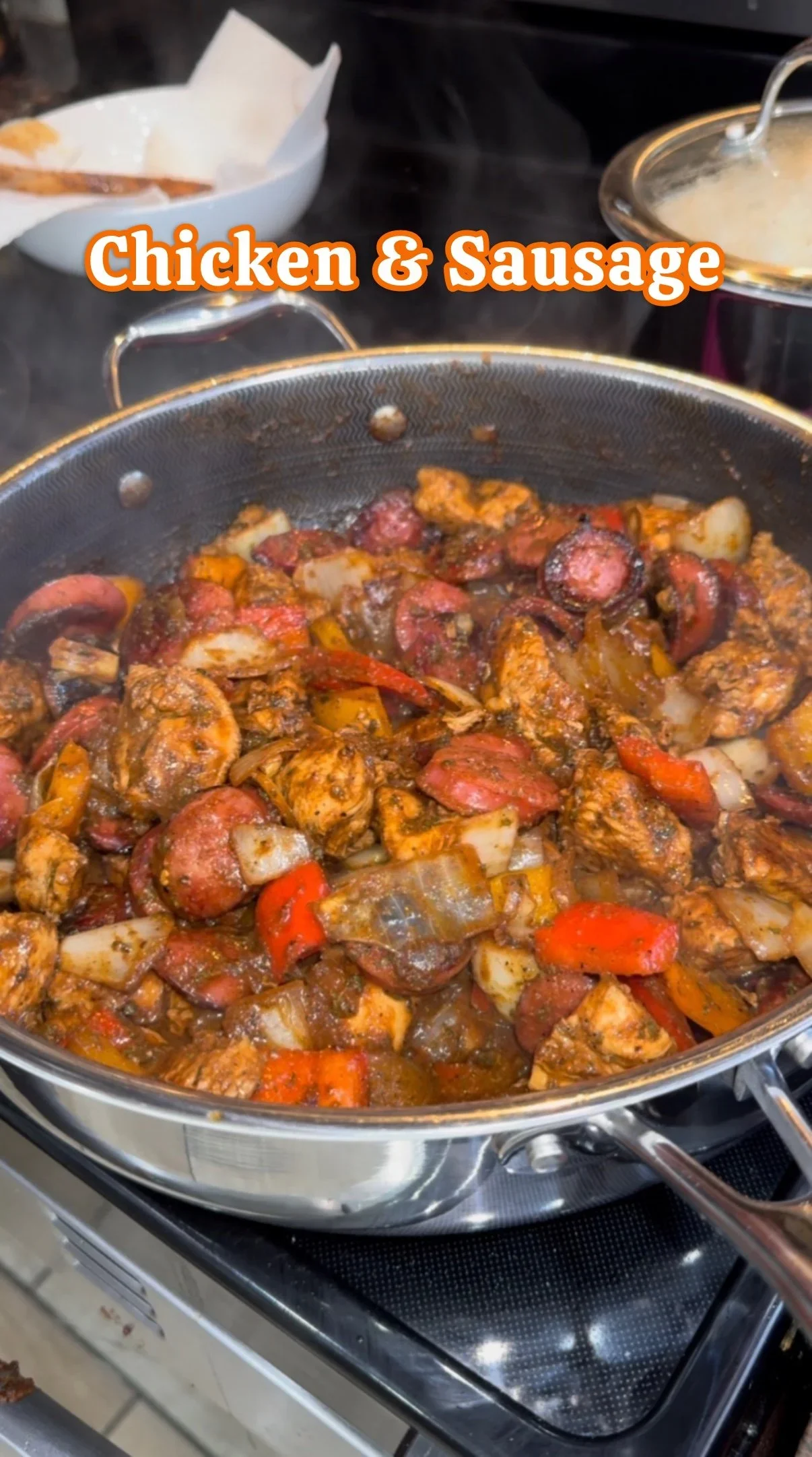 Close-up of a cooking pan filled with chicken, sausage, and mixed vegetables in a sauce or seasoning. In the background, a bowl with paper towels and a pot with liquid, possibly gravy or sauce, are visible.