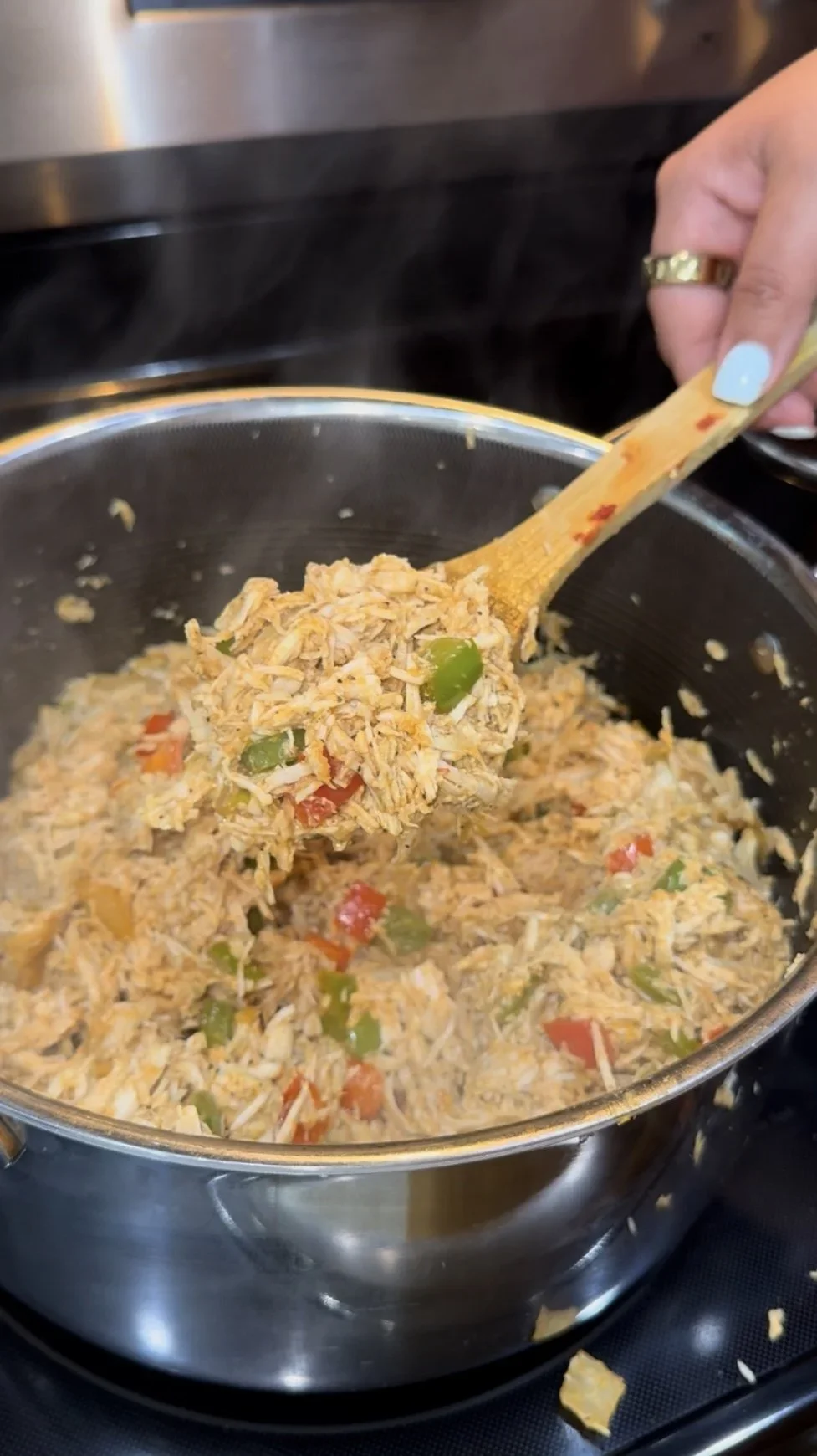 Hand stirring cooked shredded chicken with diced green and red bell peppers in a pot on the stove.