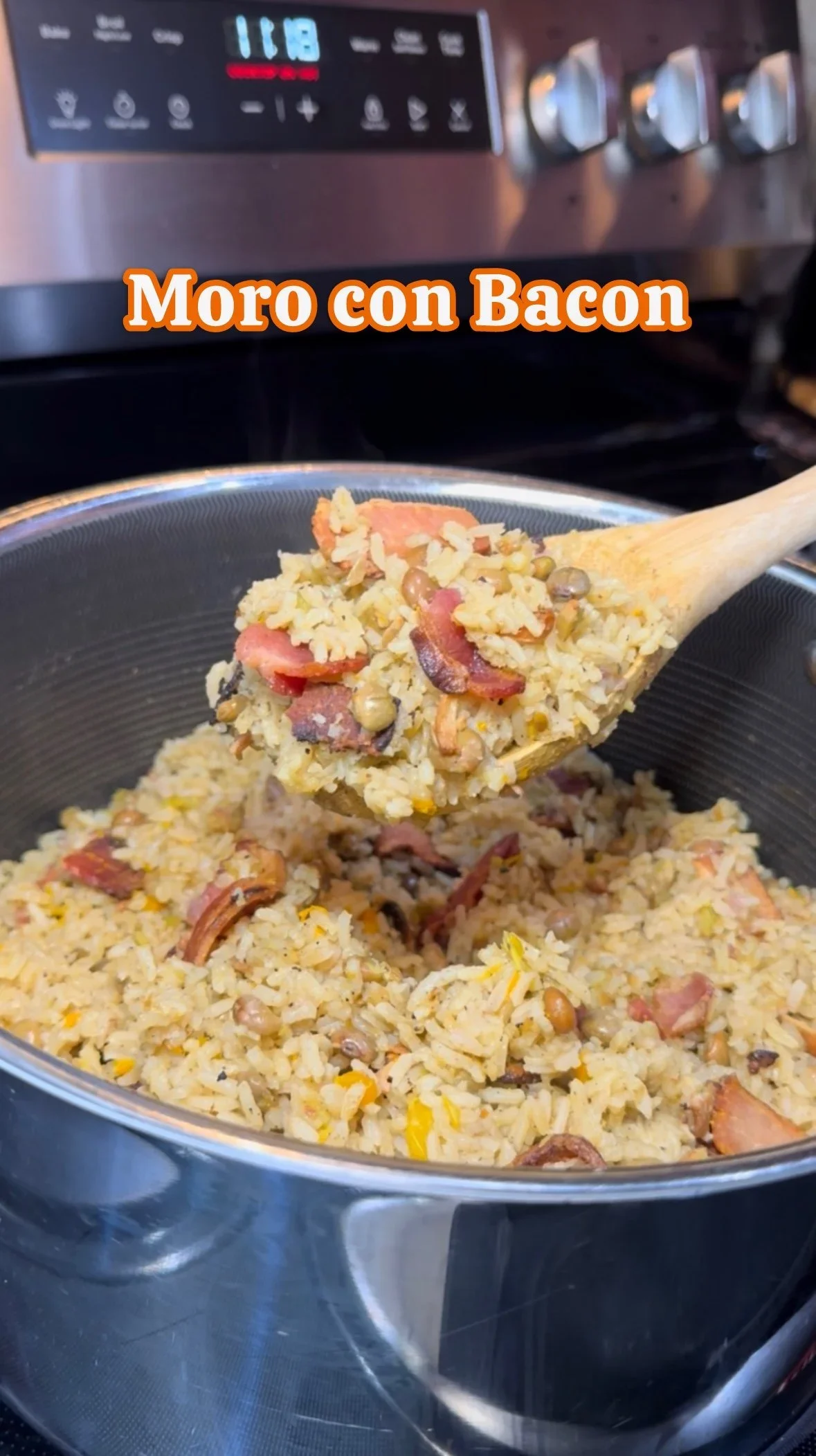 Close-up of a pot of rice with bacon and lentils, being stirred with a wooden spoon in a kitchen, labeled "Moro con Bacon".