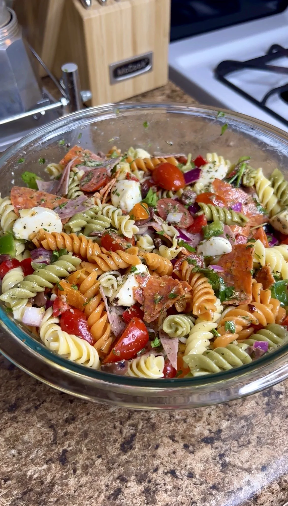 A glass bowl filled with colorful tri-colored rotini pasta salad with cherry tomatoes, red onions, herbs, pepperoni, salami and mozzarella cheese on a kitchen countertop.