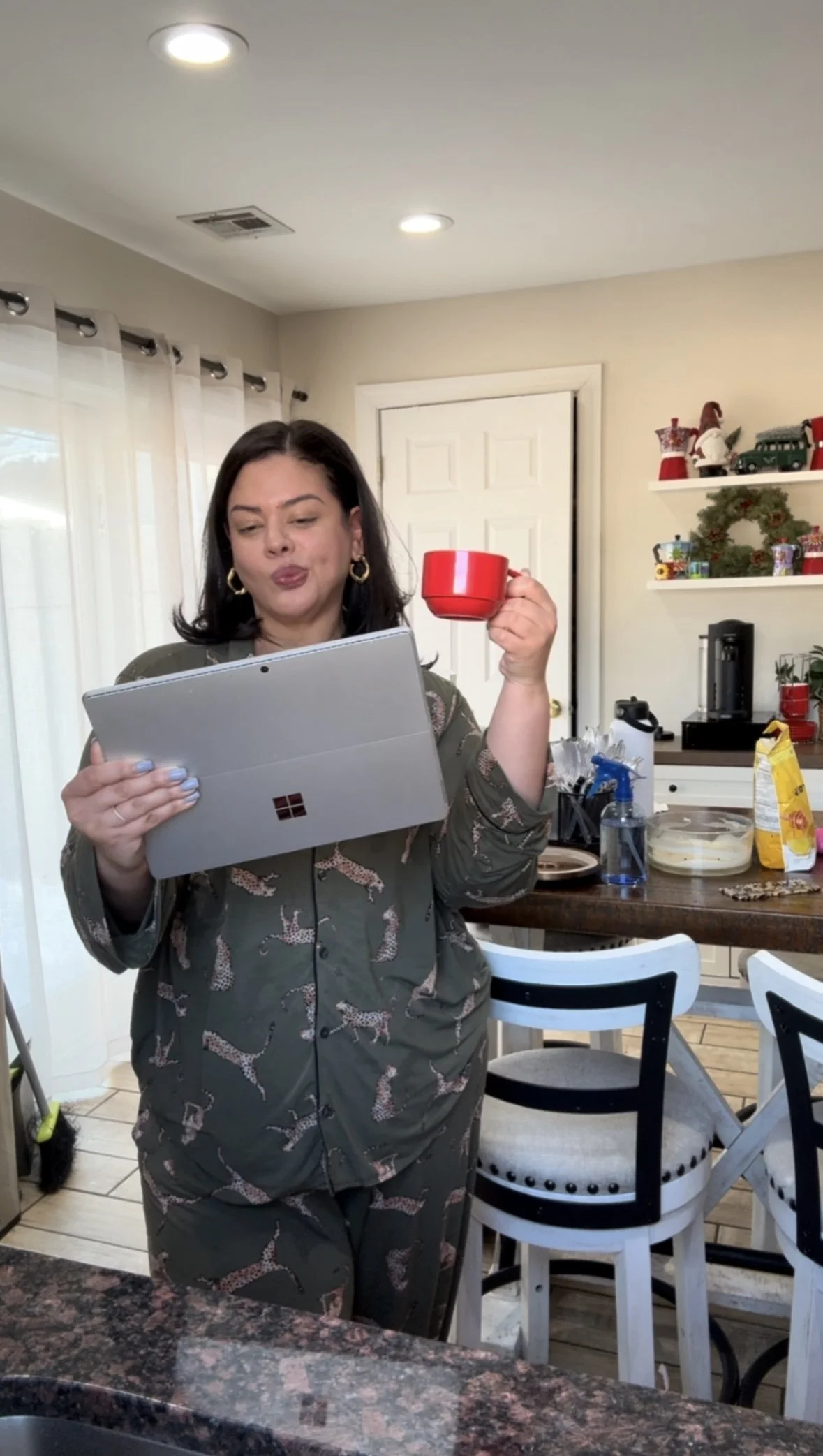 A woman standing in a kitchen, looking at a tablet in her hand while holding a red mug, with holiday decorations and kitchen items visible in the background.