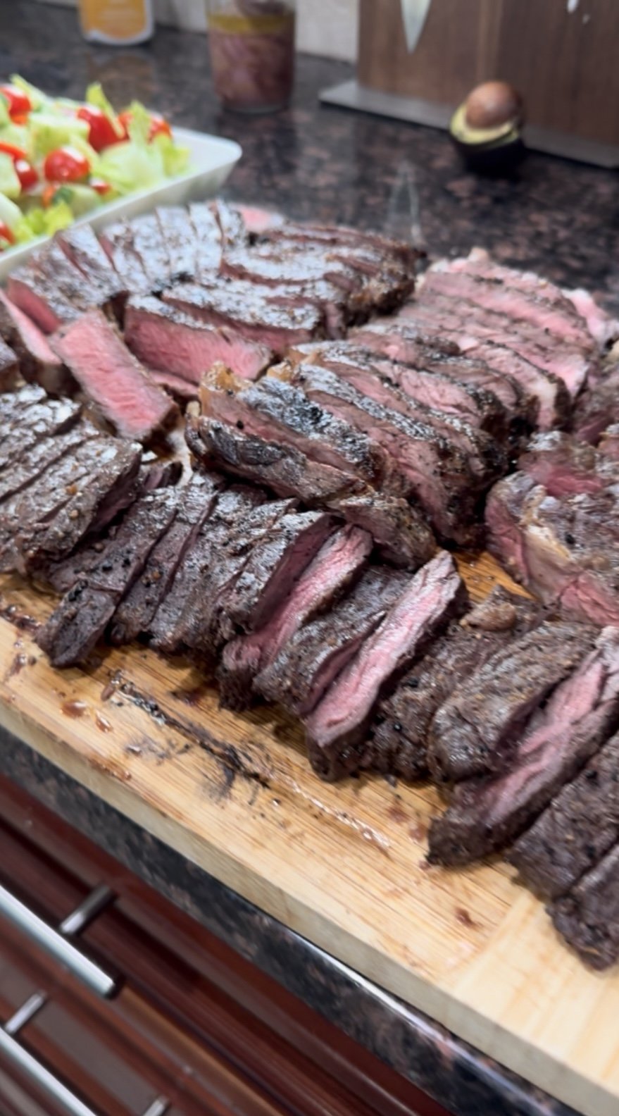 Sliced cooked steak on a wooden cutting board with salad in the background.