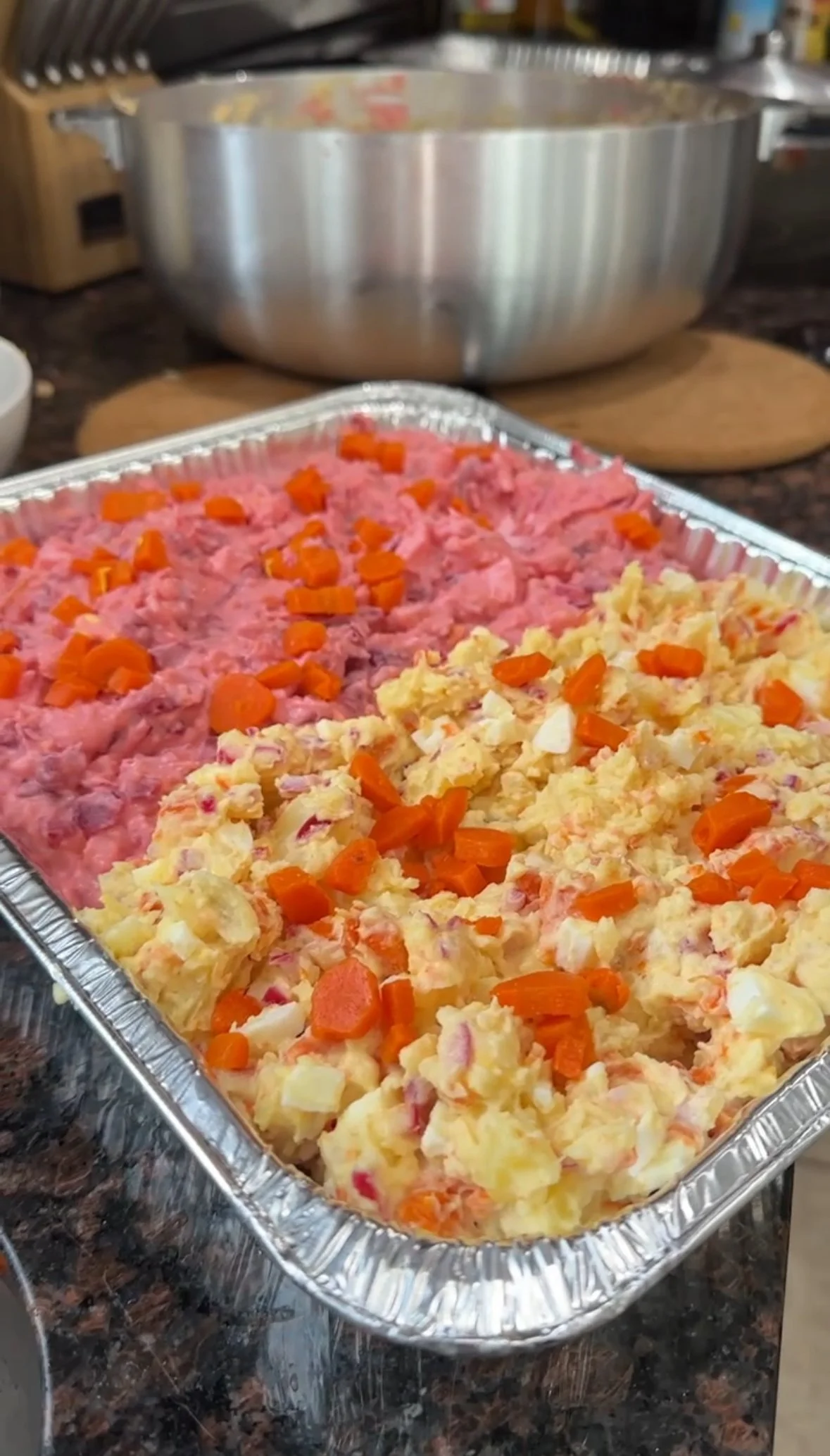 A tray of dominican potato salad with chopped carrots and a dish of pink creamy salad from beets, garnished with chopped carrots, on a kitchen counter.