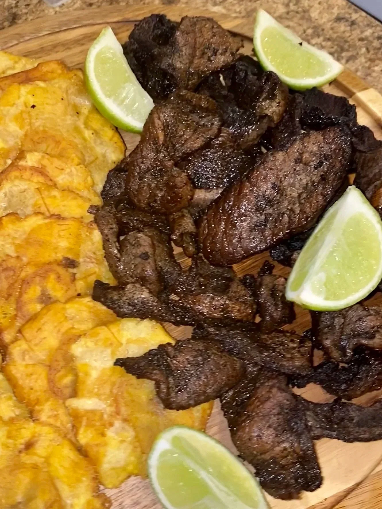Fried plantains, cooked beef steaks, and lime wedges on a wooden serving board