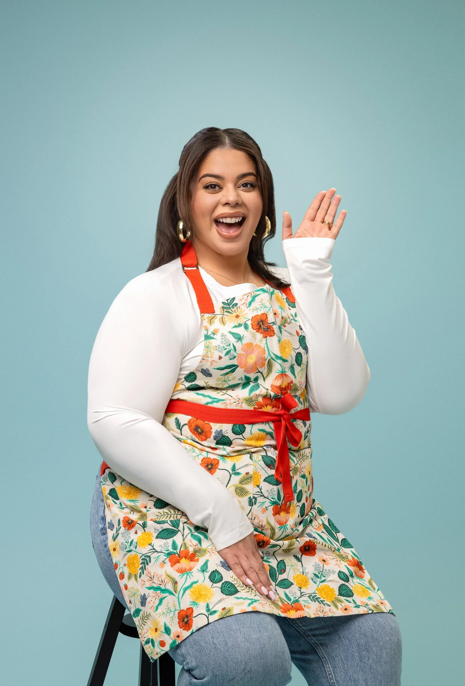 Michelle, a woman with dark hair, wearing large hoop earrings and a floral apron over a white long-sleeve shirt, sitting and smiling while waving at the camera.