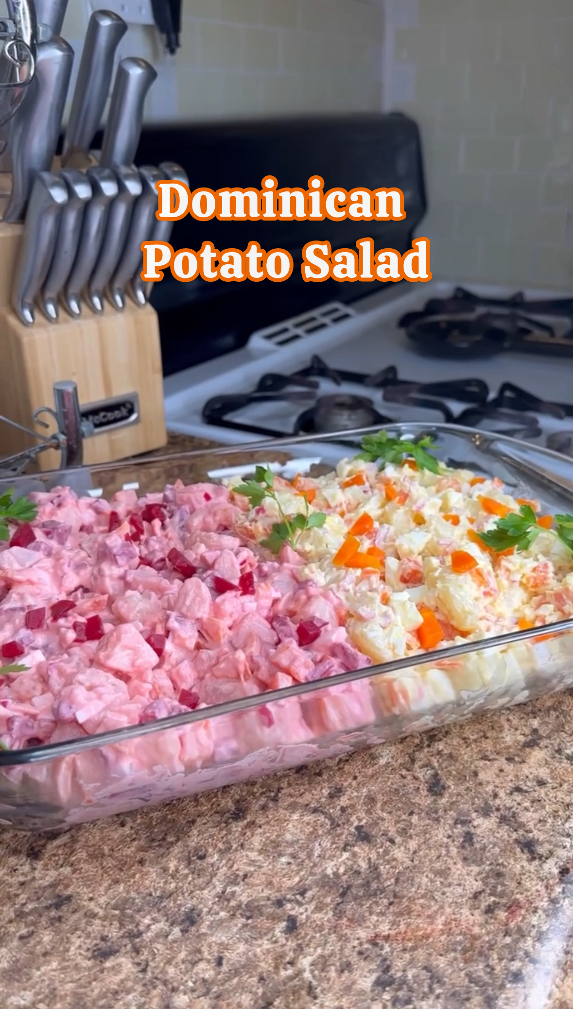 A glass dish with a serving of Dominican potato salad topped with parsley, sitting on a kitchen counter, with a knife block and stove in the background.