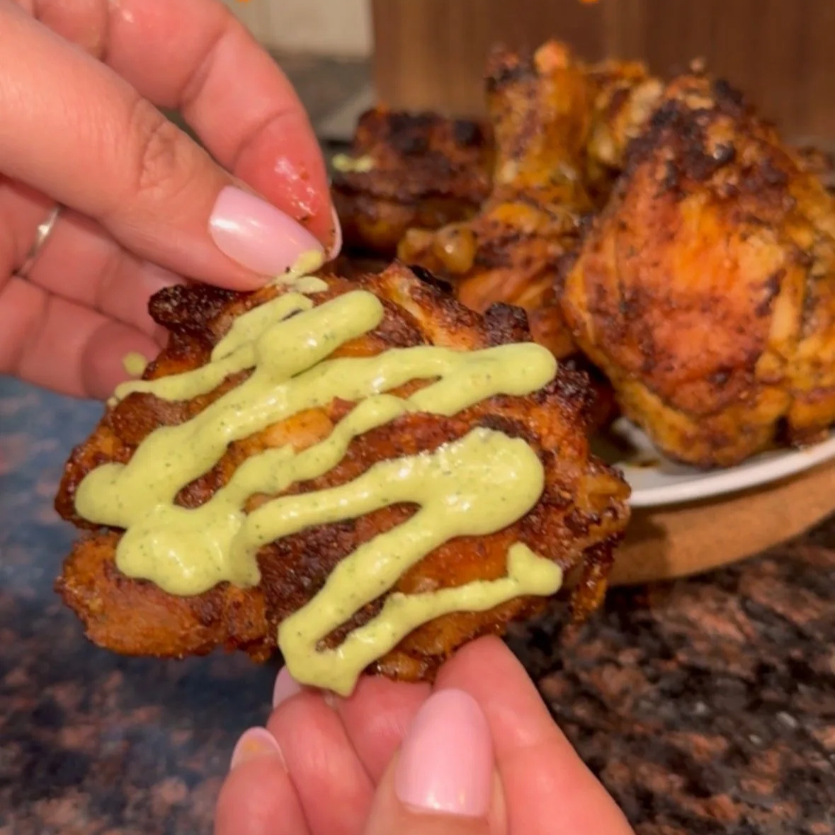 Close-up of someone holding a piece of baked chicken drizzled with creamy green sauce, with whole baked chicken pieces on a plate in the background.
