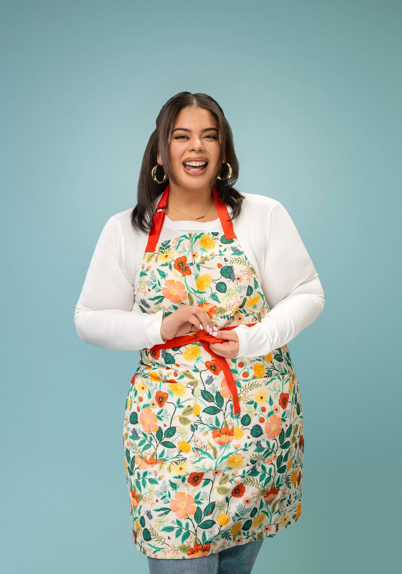 A woman with dark hair and gold hoop earrings wearing a white long-sleeve shirt and a colorful floral apron, smiling and holding the apron tie against a light blue background.