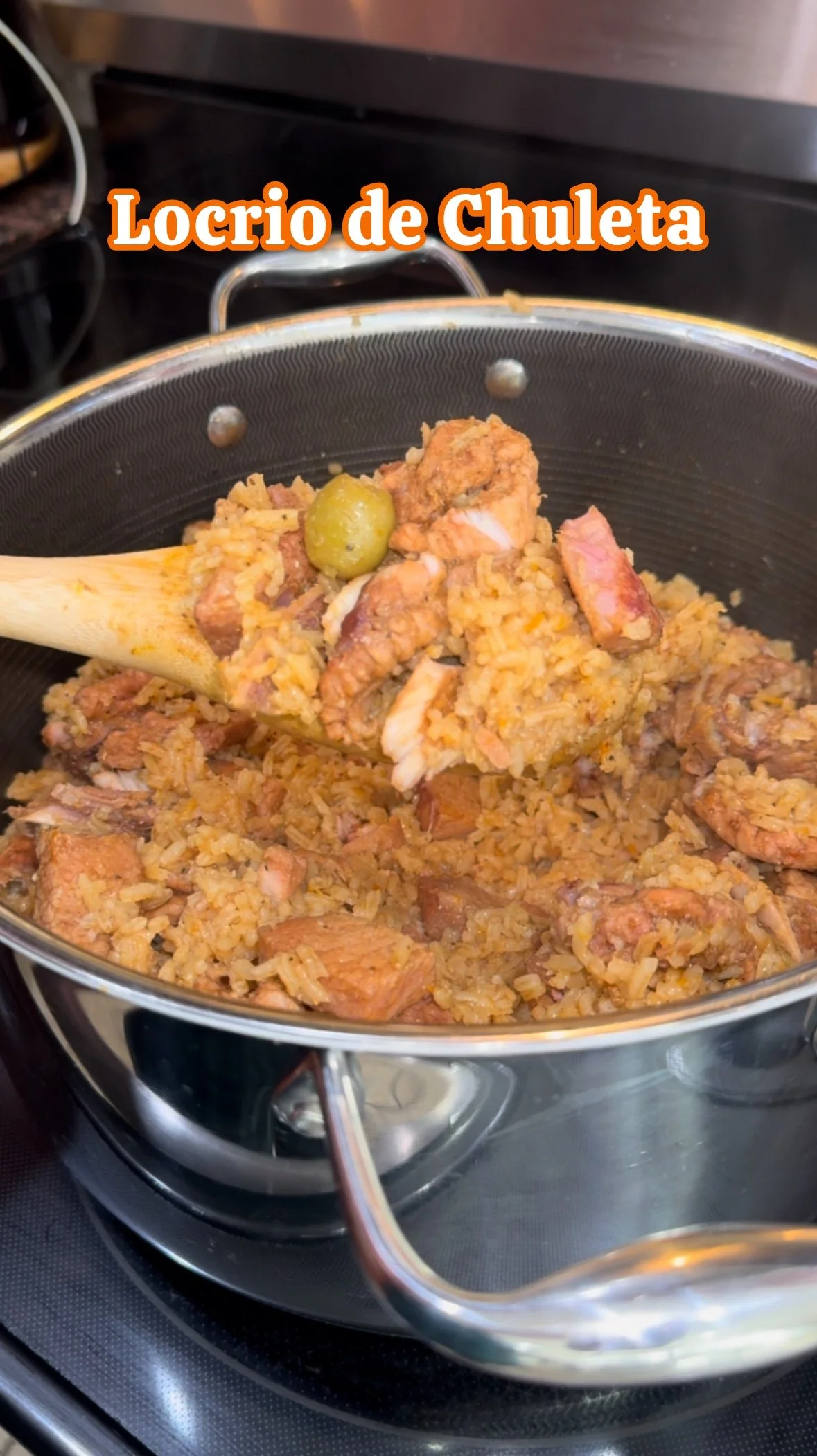 A pot of Mexican rice with chunks of pork chop and a green olive, with a wooden spatula scooping some rice, labeled 'Locrio de Chuleta'.
