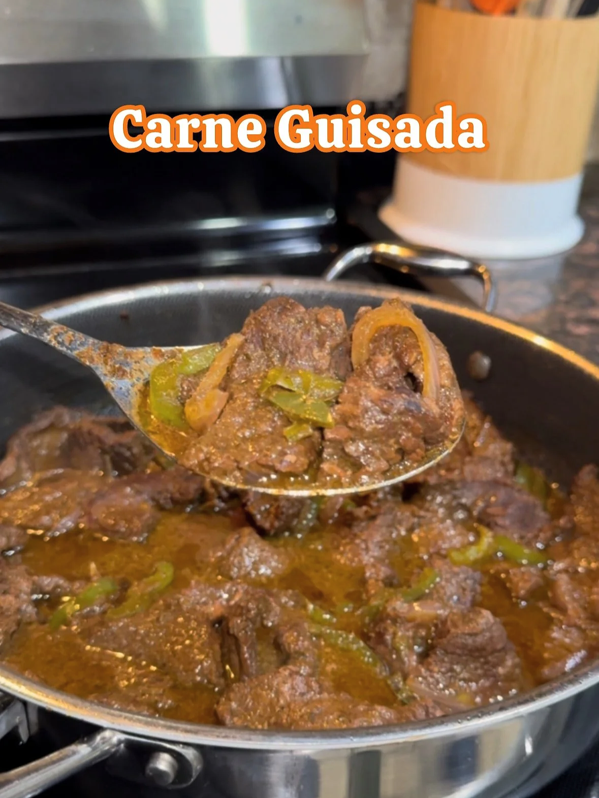A spoonful of cooked carne guisada, a beef stew with green peppers and onions, held above a skillet of the same dish.