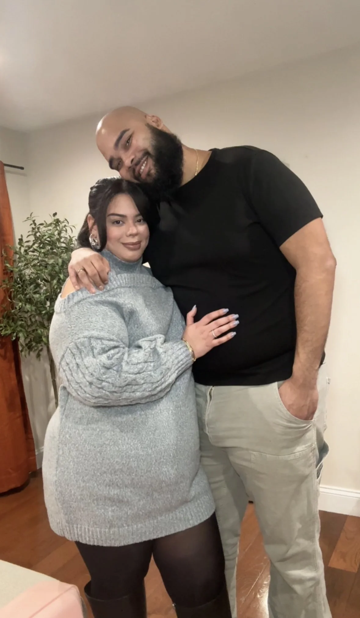 A man and woman are standing close together indoors, smiling at the camera. The man has a bald head, beard, and is wearing a black t-shirt and beige pants. The woman has dark hair, is wearing a gray sweater dress, black tights, and earrings. They appear to be in a cozy room with a wooden floor, a green plant, and light-colored walls.