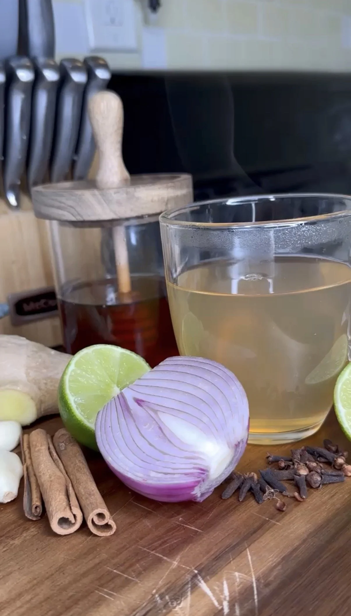 A glass of cold-buster tea, a jar of honey with a wooden honey dipper, fresh lime halves, a halved red onion, cinnamon sticks, and cloves on a wooden surface.