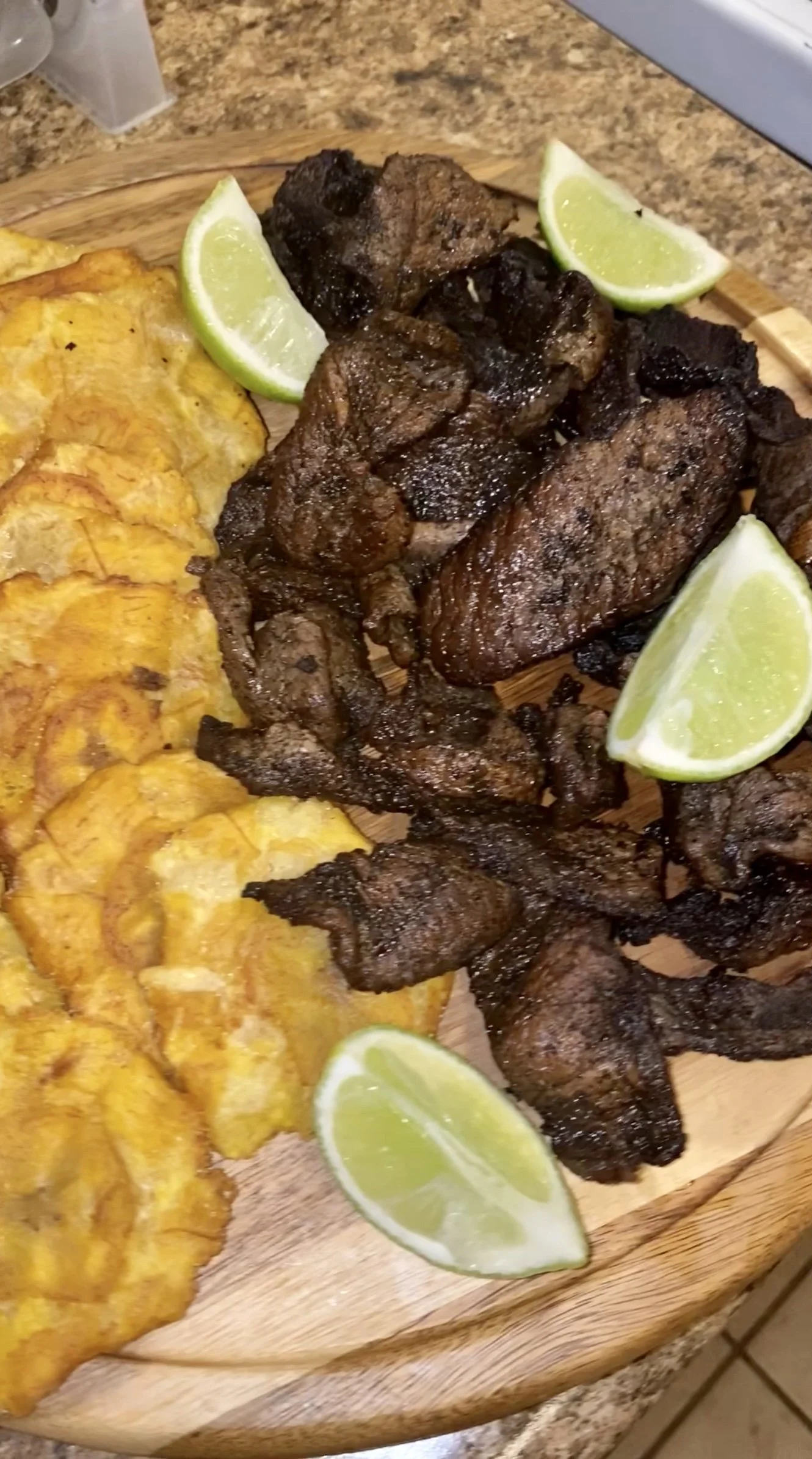 Fried plantain/tostones with fried beef chunks, lime wedges on a wooden platter.