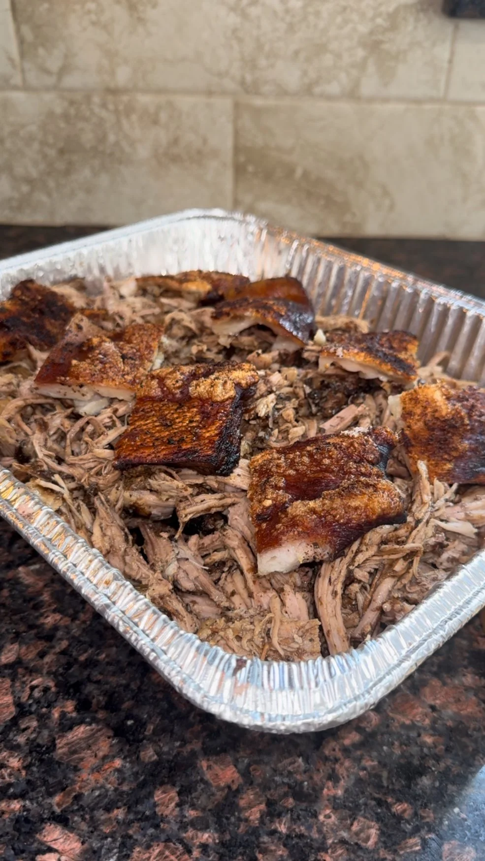Cooked pulled pork with crispy pork belly pieces in a foil tray on a dark granite countertop.