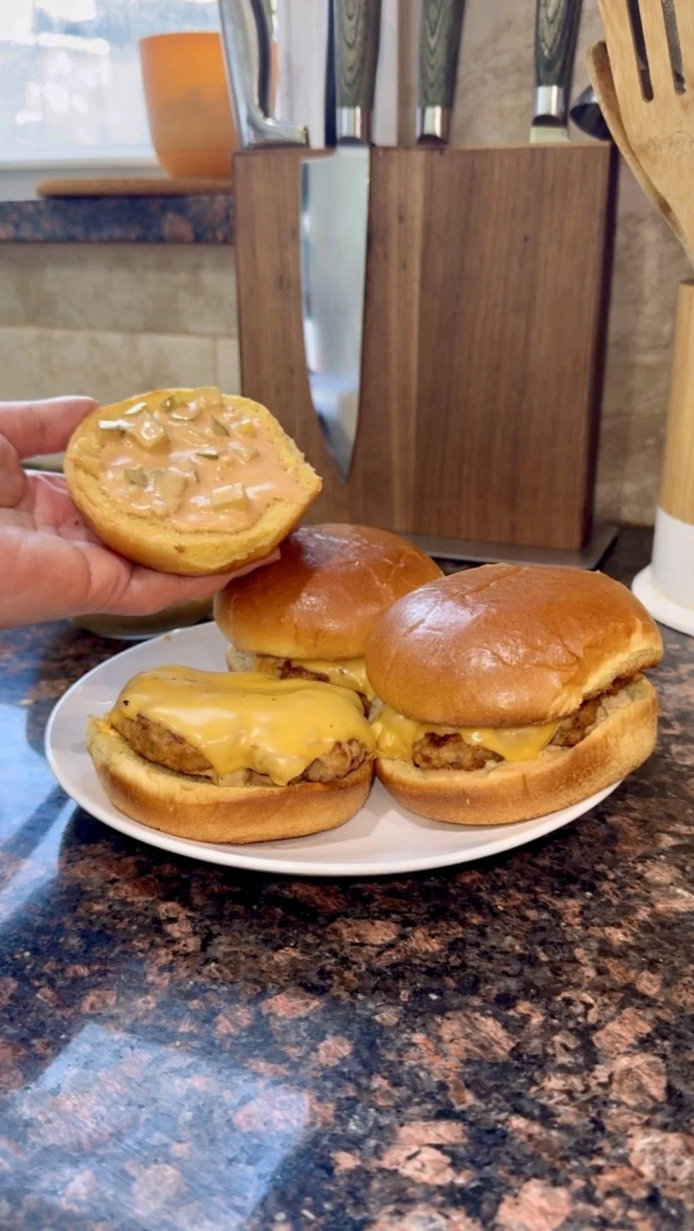 Three ground chicken burgers on a white plate with a hand holding a potato bun displaying a creamy sauce