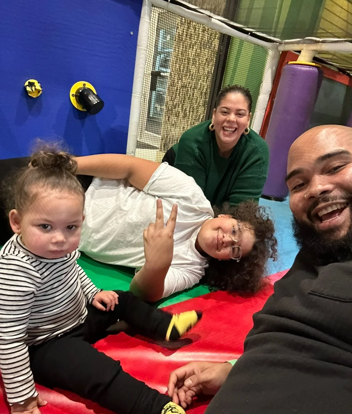 Group of four people, two children and two adults, taking a selfie at a colorful indoor play area. One child with curly hair and glasses is lying on a red and green padded mat, making a peace sign. The other child, with a serious expression, is sitting cross-legged. One woman with dark hair tied back and another man with a beard are smiling at the camera.