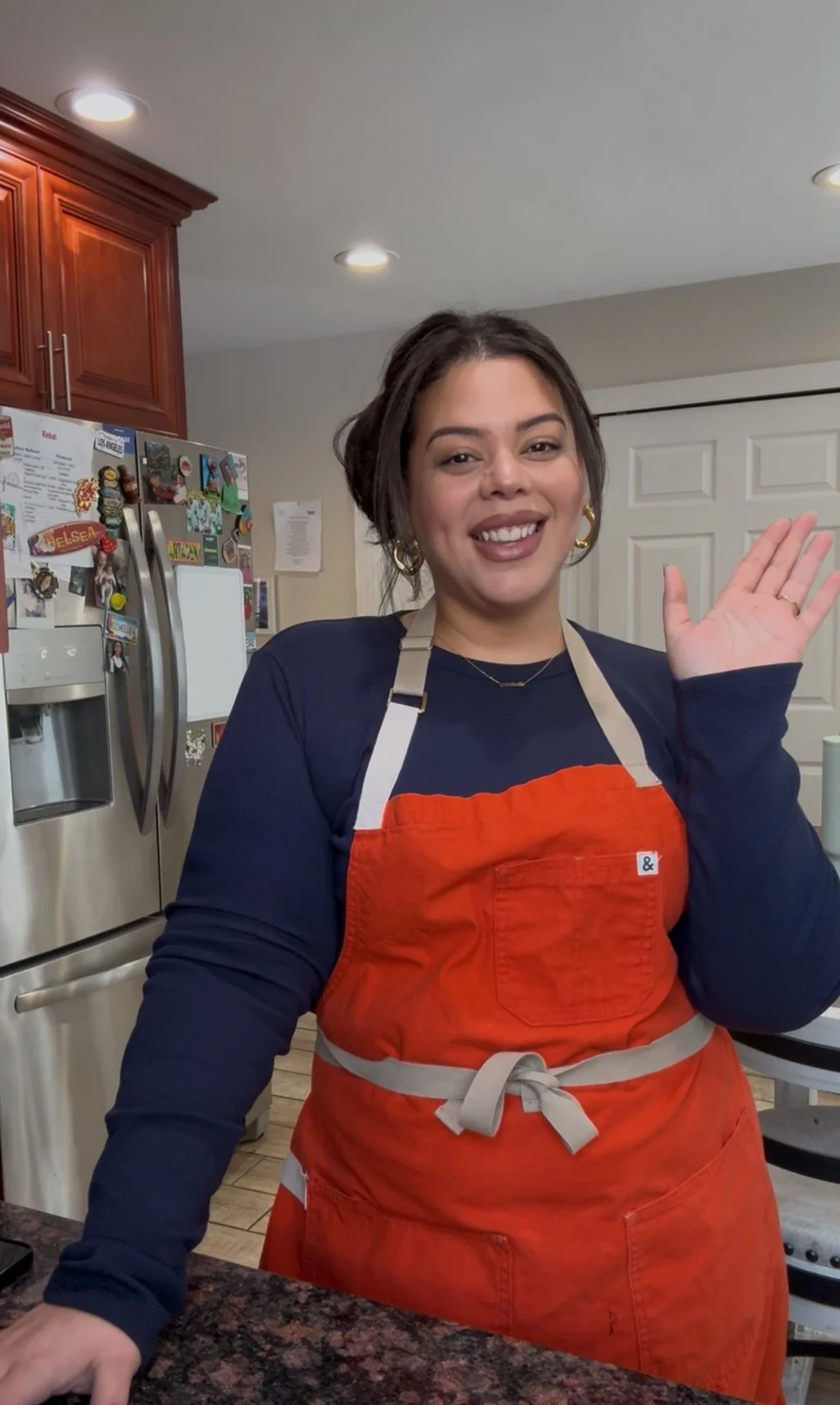 Michelle, a smiling woman with dark hair in a bun, wearing a navy blue shirt and orange apron, waving in a kitchen with stainless steel refrigerator and wooden cabinets.