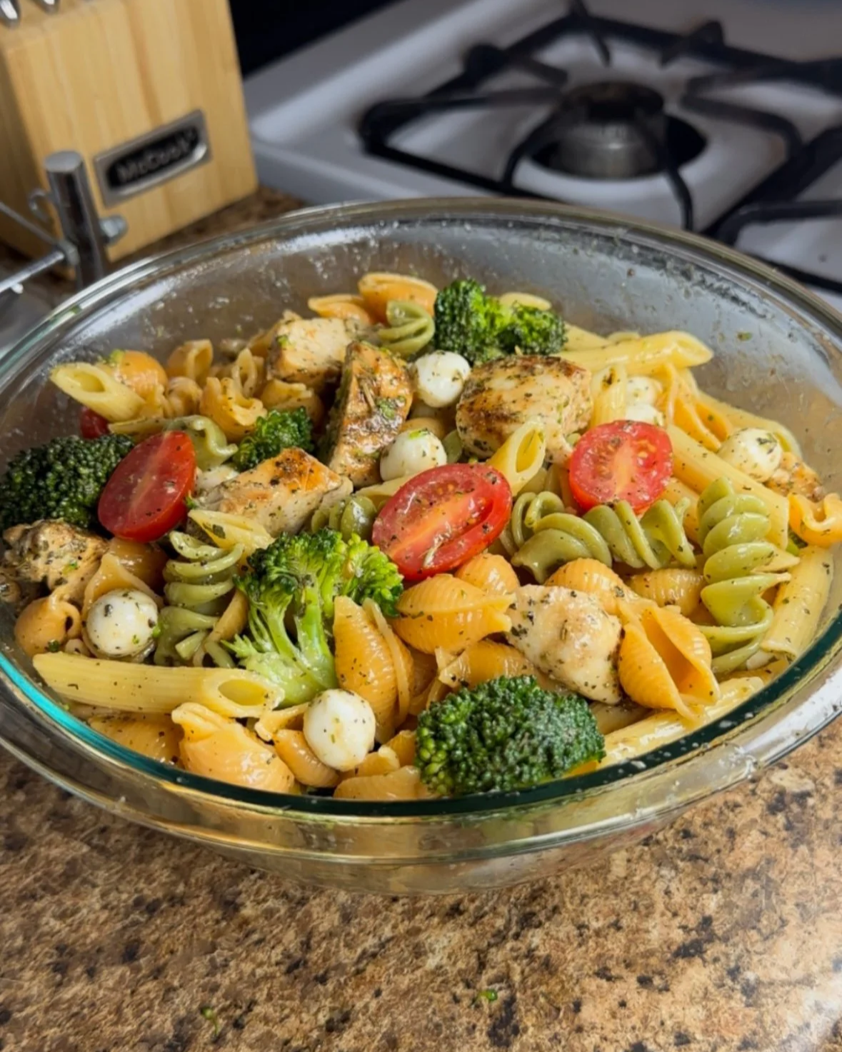 A glass baking dish filled with pasta, cherry tomatoes, broccoli, mozzarella balls, grilled chicken pieces, and green pasta, all seasoned with herbs, sitting on a brown countertop in a kitchen.