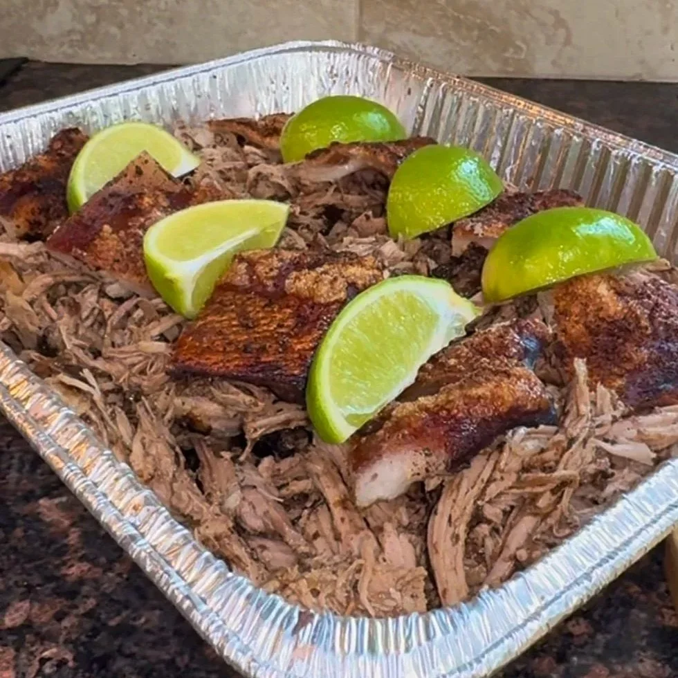 Oven roasted pork shoulder garnished with lime wedges in a foil tray.