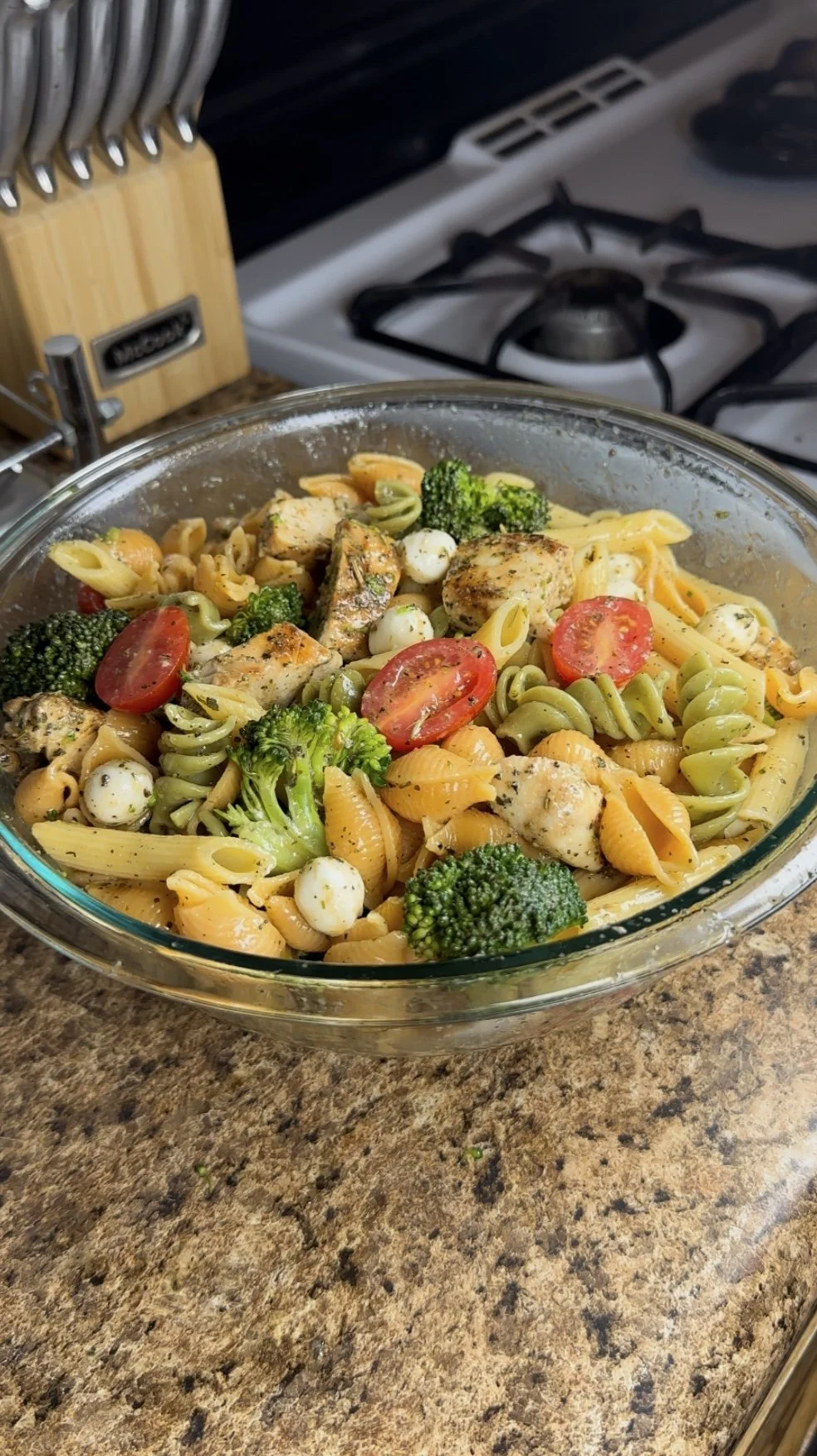 A glass baking dish filled with cooked pasta, broccoli, cherry tomatoes, chicken pieces, and mozzarella balls on a kitchen countertop.