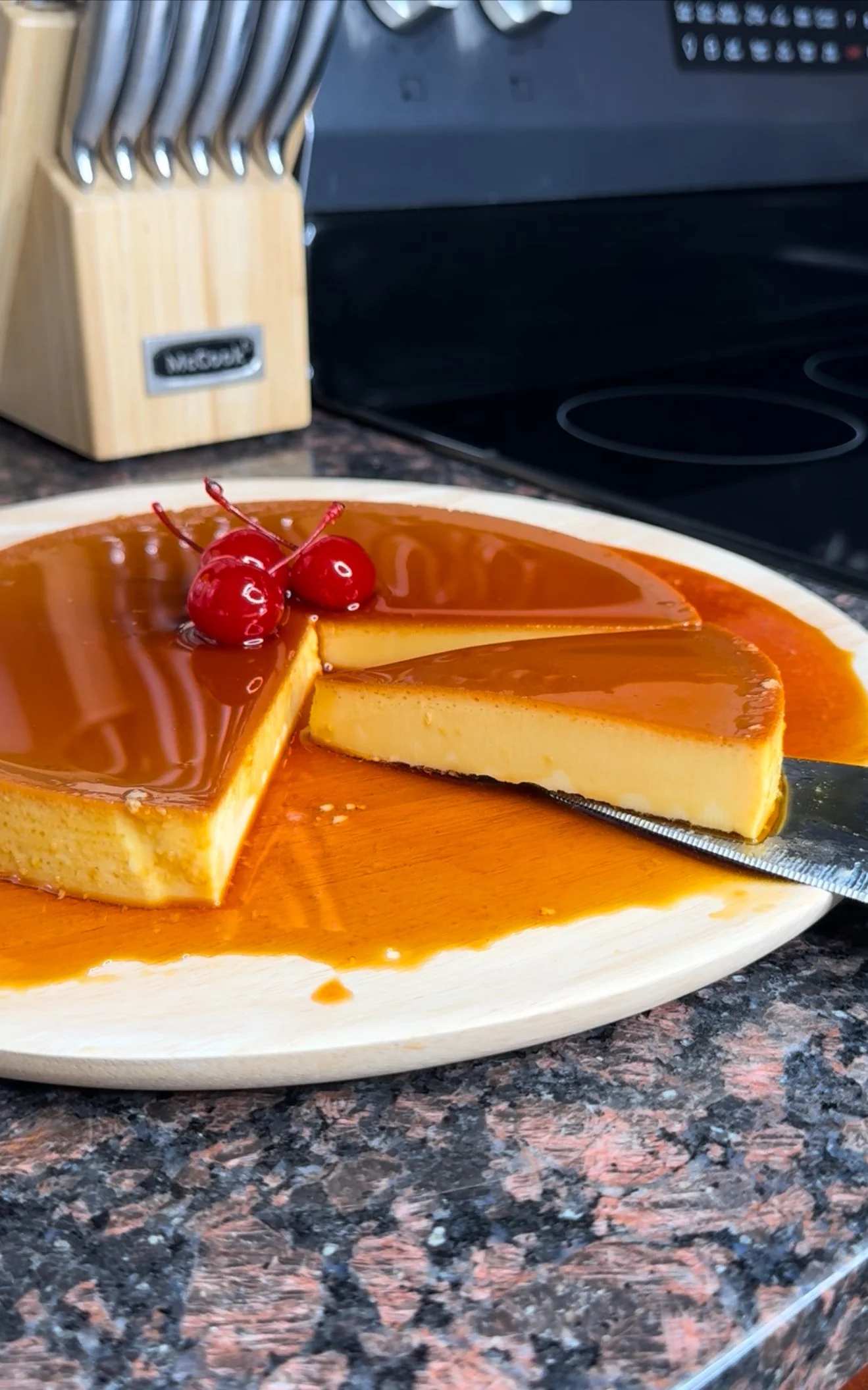 A caramel flan with cherries on top on a white plate on a kitchen countertop.