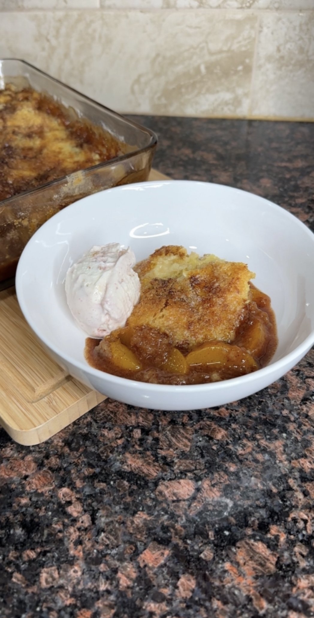 Peach cobbler with a scoop of vanilla ice cream in a white bowl on a dark granite countertop.