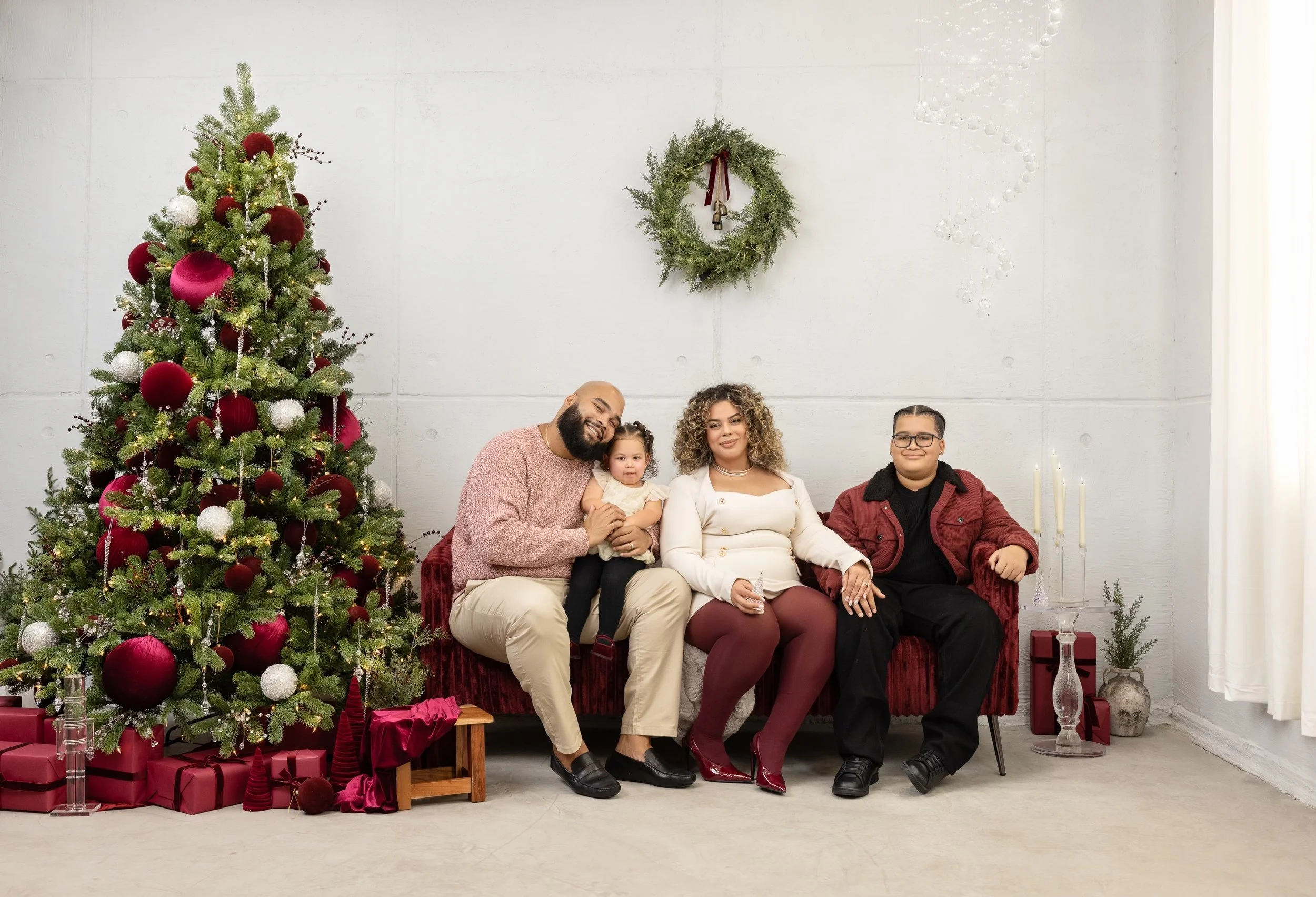 Michelle's family, sitting on a red sofa in a festive photo studio with a Christmas tree decorated with red and white ornaments, presents underneath, and holiday decor, including a wreath on the wall and candles on the side table.