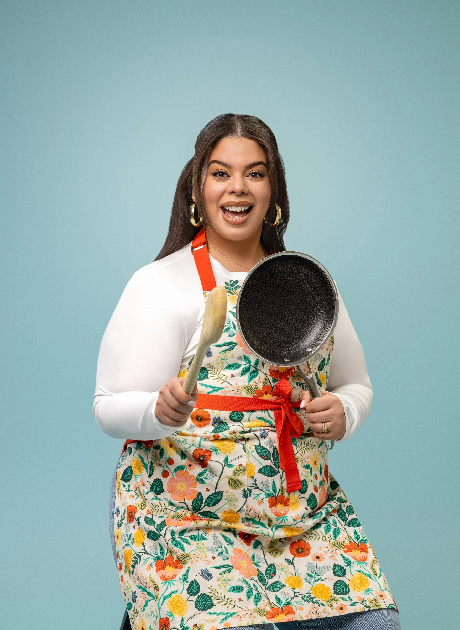 A woman wearing a floral apron, holding a frying pan and a spatula, smiling at the camera against a light blue background.
